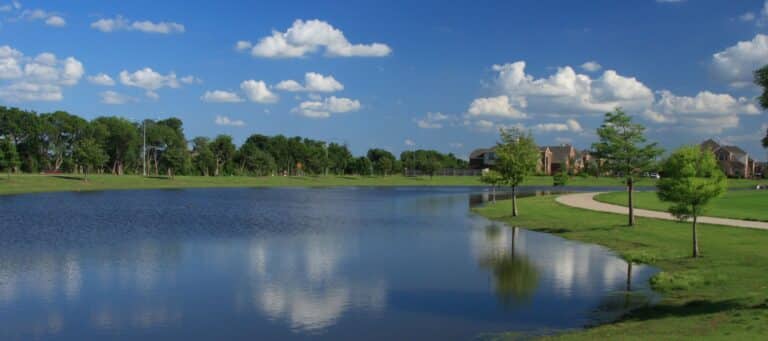 A serene pond reflects the blue sky and scattered clouds, bordered by a grassy path and trees, with houses visible in the distance