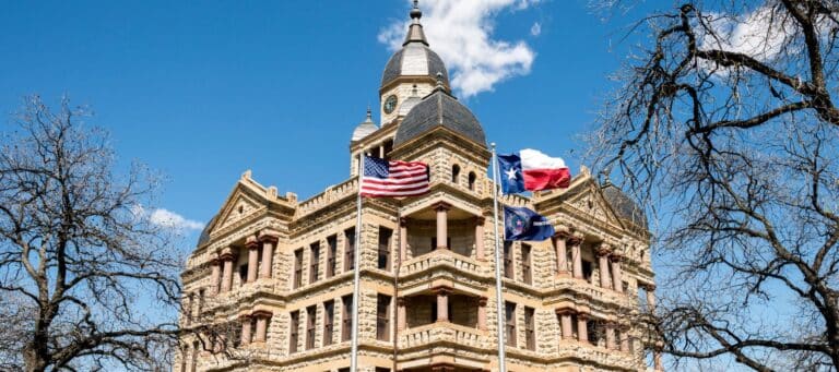 Historic courthouse building with American, Texas, and local flags waving in front, framed by bare trees and a bright blue sky