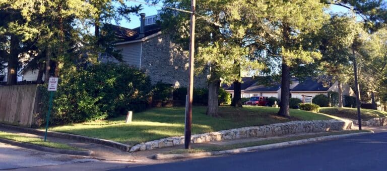 Residential neighborhood with stone retaining walls, trees, and a house partially hidden by greenery