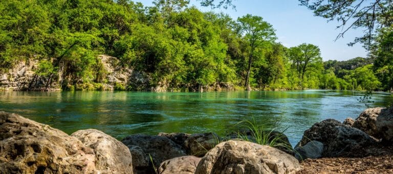 Clear blue river surrounded by greenery and rocks, flowing through a tranquil forested area