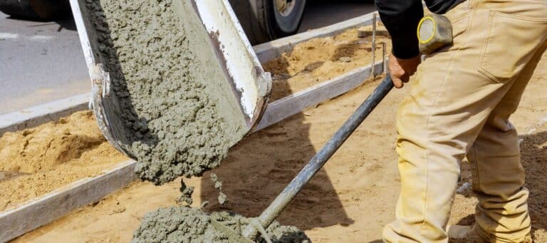 A construction worker guiding freshly poured concrete from a mixer into a framework, preparing for a sidewalk or foundation