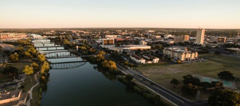 A scenic aerial view of downtown Waco, Texas, showcasing bridges over a calm river and the city's skyline at sunset
