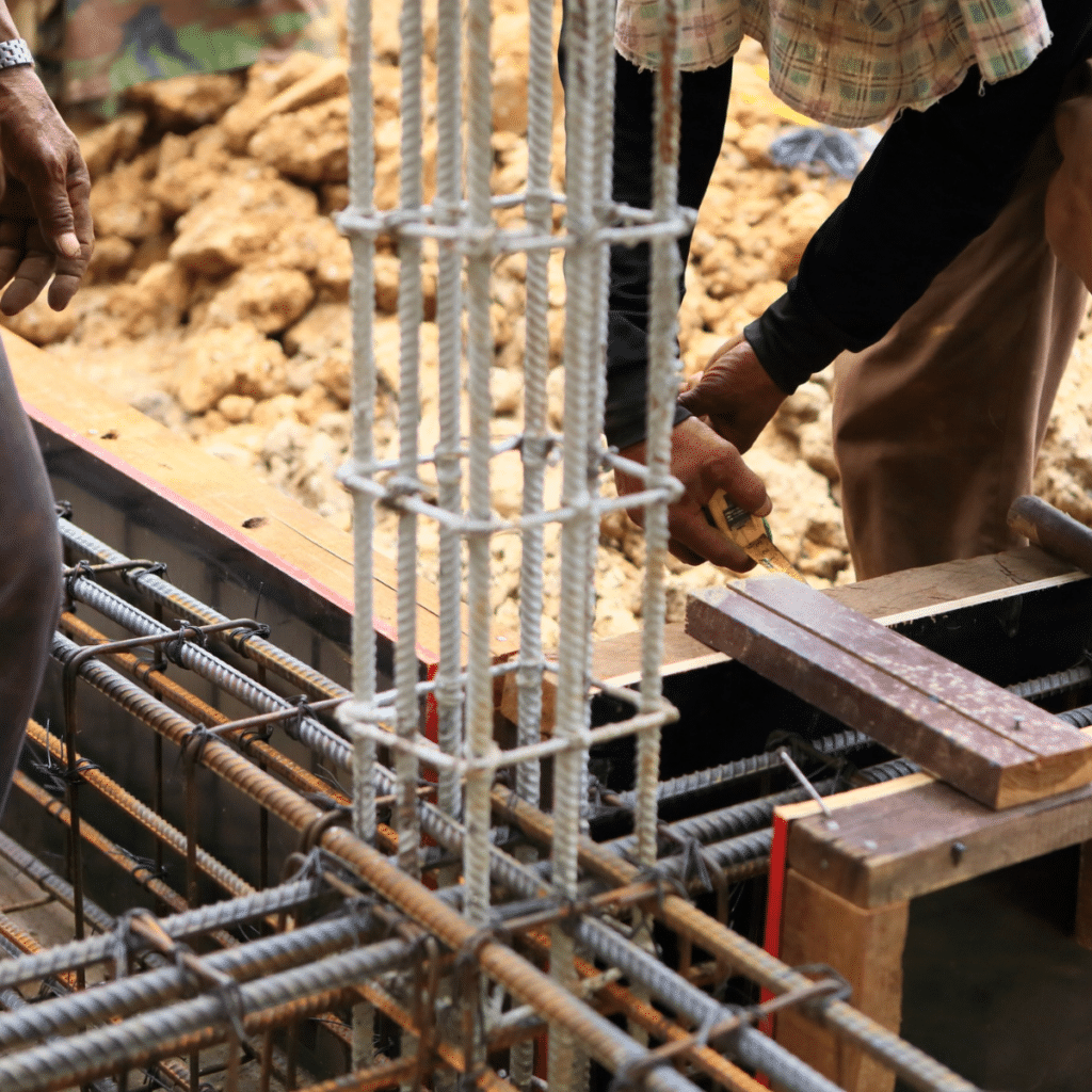 orkers assembling steel reinforcement bars (rebar) for a concrete foundation, with tools, wooden planks, and soil visible in the construction site background