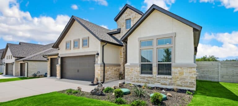 A modern suburban home with stone and stucco exterior, a neatly landscaped front yard, and a bright blue sky in the background