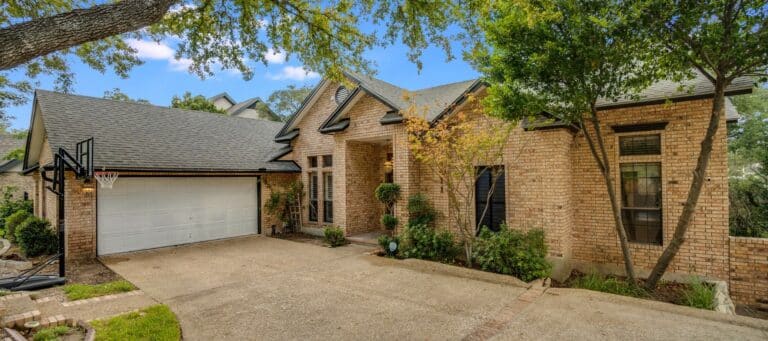 Sloped driveway leading to a two-car garage of a brick home surrounded by mature trees