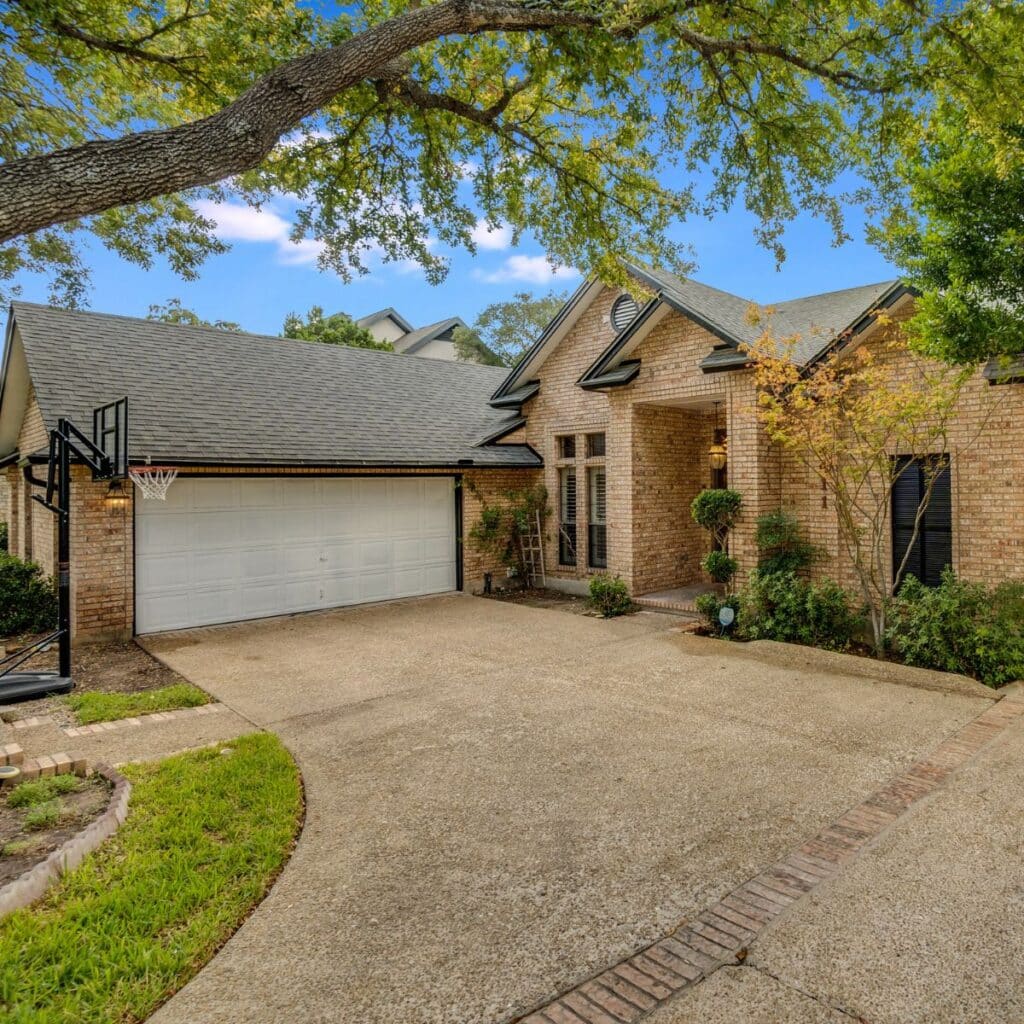 Sloped driveway leading to a two-car garage of a brick home surrounded by mature trees