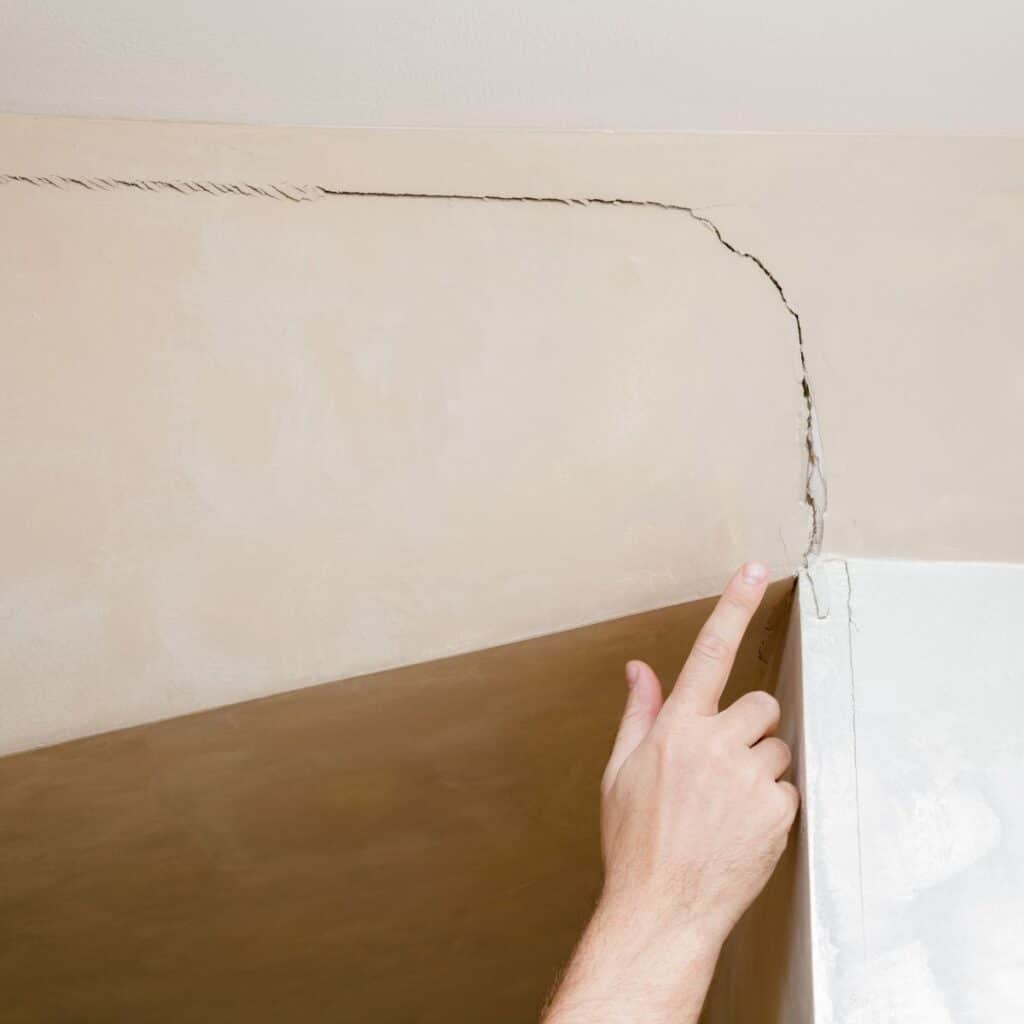 Person pointing at a large diagonal crack running across a beige interior wall, indicating possible foundation issues