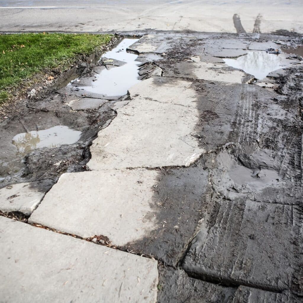Broken and uneven concrete sidewalk with muddy patches and standing water