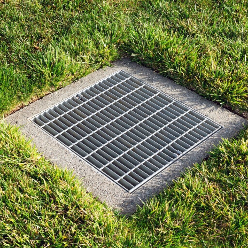 Metal surface drain grate embedded in a concrete square surrounded by grass