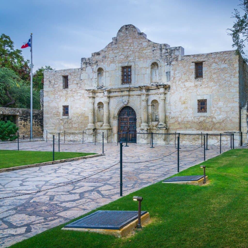 Front view of the historic Alamo mission in San Antonio, Texas, with its iconic limestone façade and a Texas flag flying on the left
