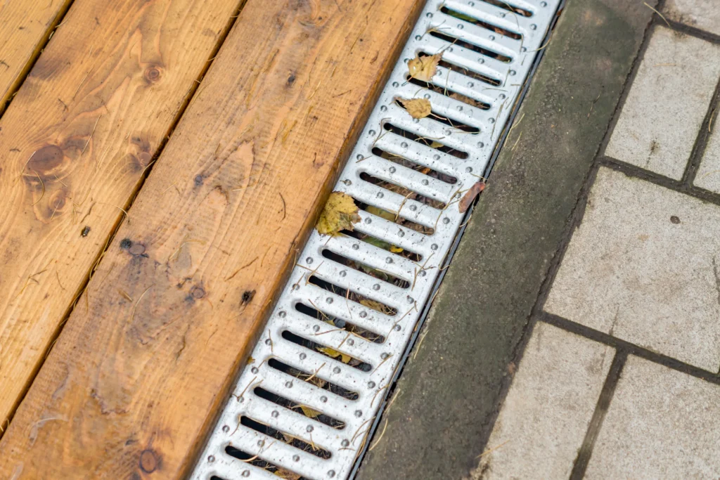 Metal drain grate installed between a wood deck and concrete walkway, channeling water away from the foundation.
