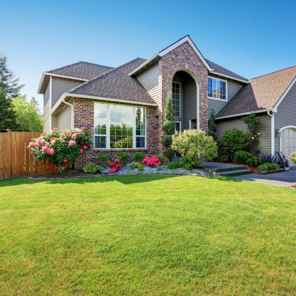 A large, two-story house with a brick and siding exterior and an arched front entry is surrounded by a green lawn and blooming pink flowers