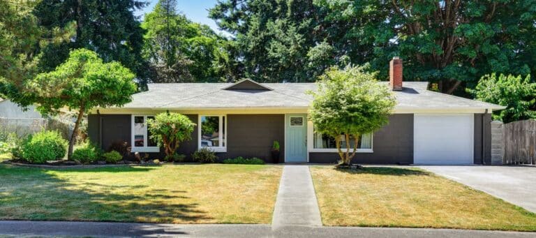 Single-story gray house with a front lawn, garage, and large trees in the background