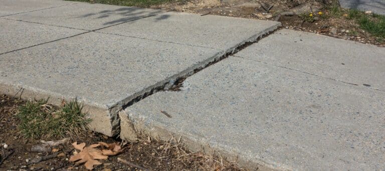 A cracked concrete sidewalk slab is significantly raised above the adjacent slab, creating a large, uneven gap