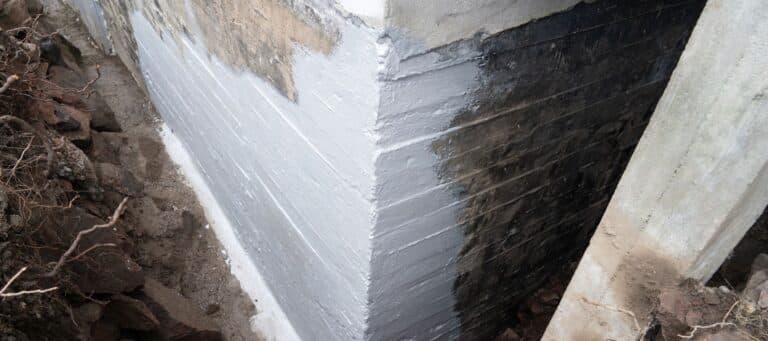 The corner of a building's foundation is seen, partially coated with a new grey sealant, with exposed earth and brick around it