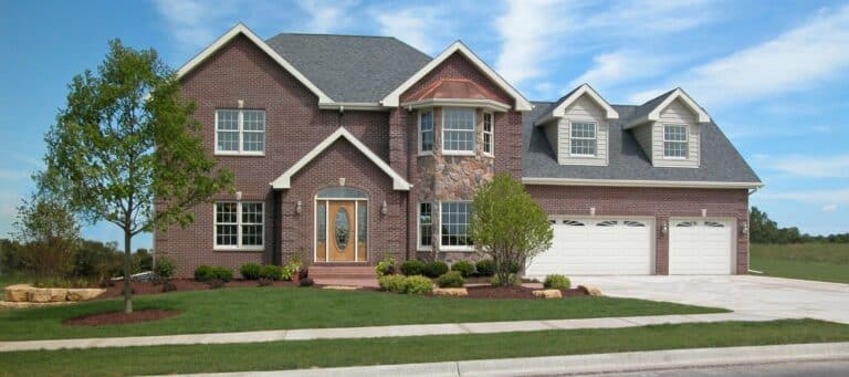 A grand two-story red brick house features a light-colored stone bay window, two white garage doors, and a green lawn under a bright, cloudy sky