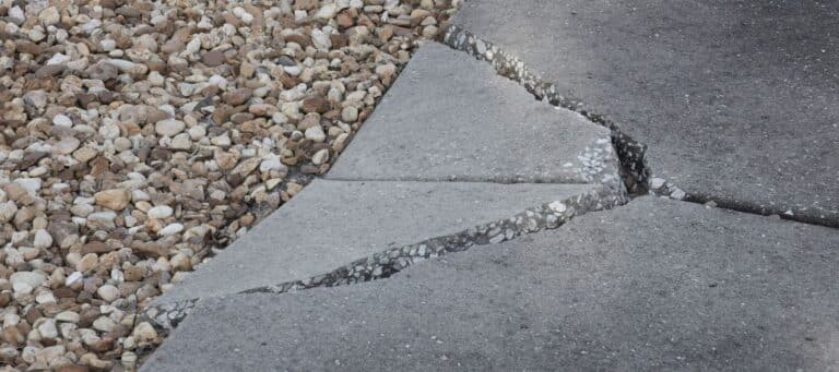 A cracked and uneven concrete sidewalk is shown next to a patch of light-colored gravel