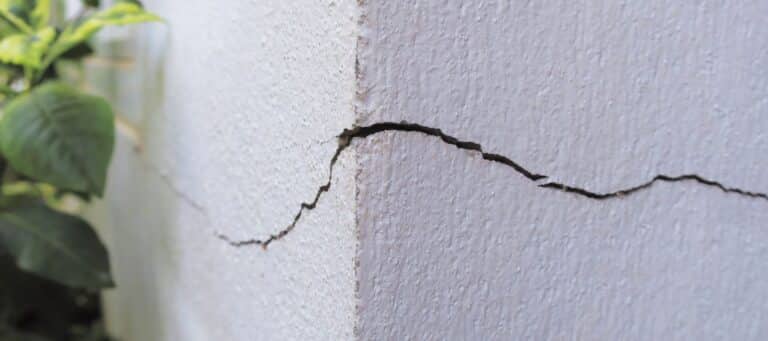 A deep, jagged crack is visible on the exterior corner of a textured white wall next to green foliage