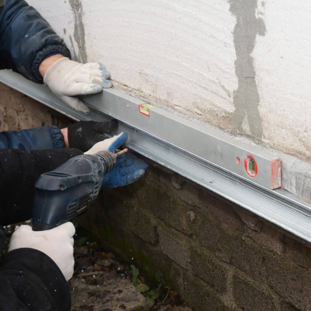 Worker using a drill and level tool to secure metal framing along a foundation wall