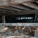 Exposed wooden beams and supports in a crawl space with debris on the ground