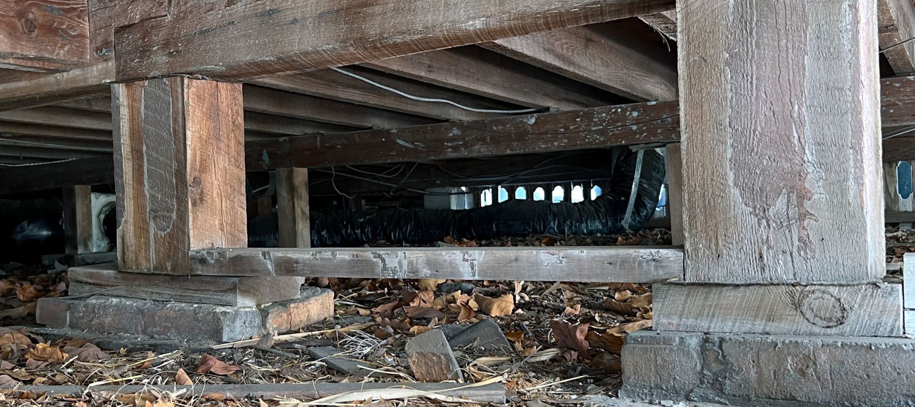 Exposed wooden beams and supports in a crawl space with debris on the ground