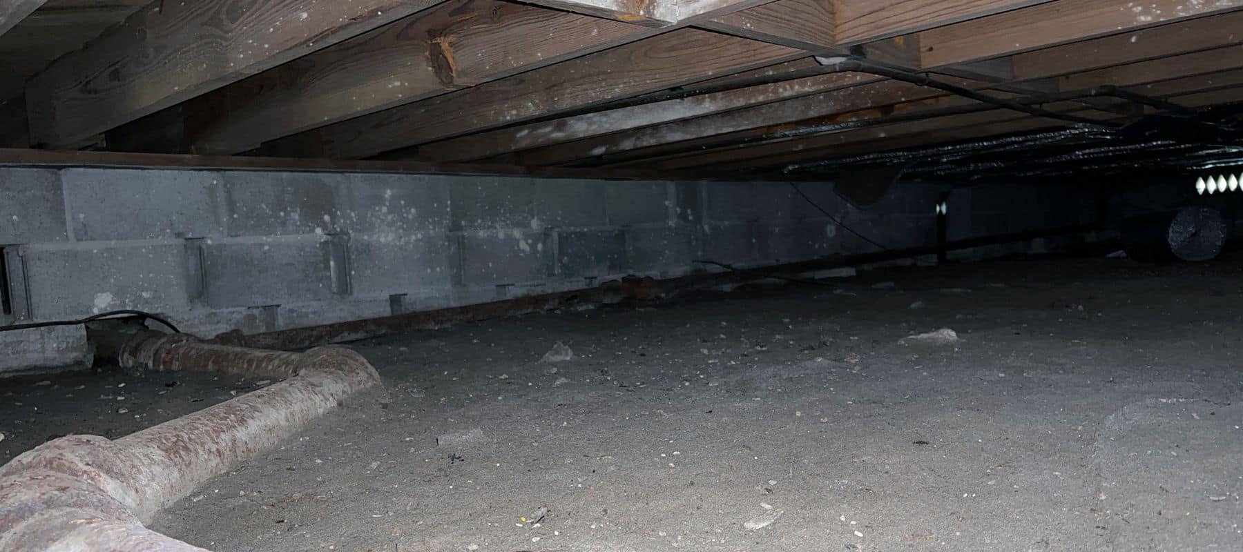 Dark crawl space under a house with exposed pipes and concrete block walls