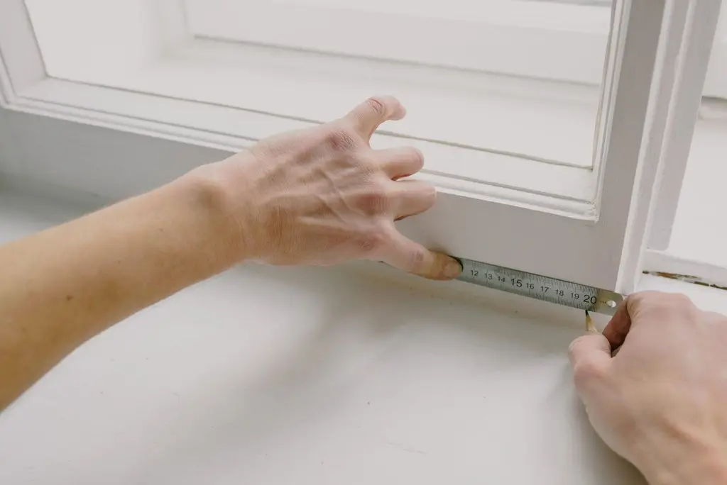 Hands using a small metal ruler to measure a triangular gap that has opened above a white window frame, indicating the wall has shifted.